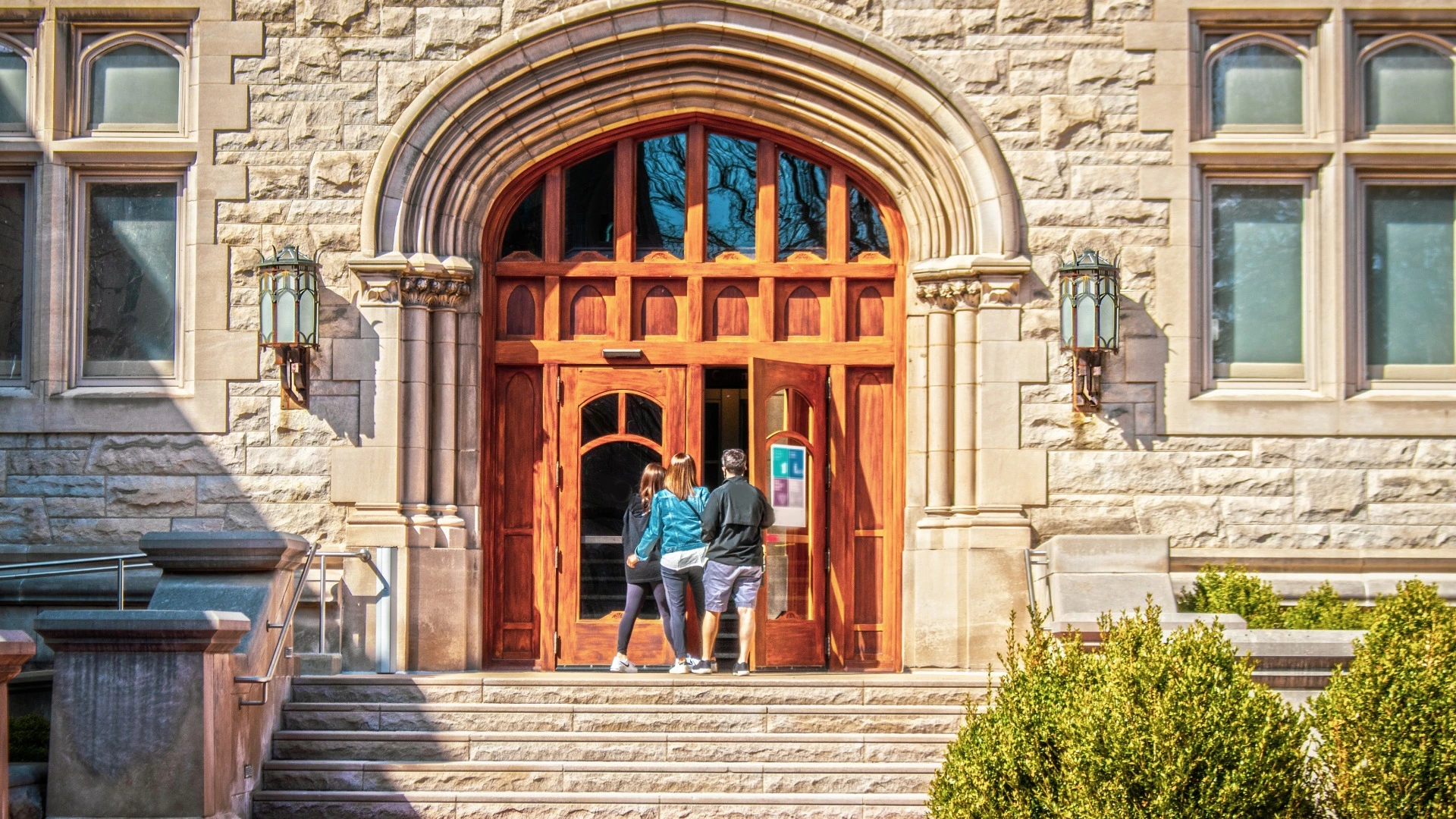 Parents and female student entering large arched doors of University building flanked by metal gothic lamps.